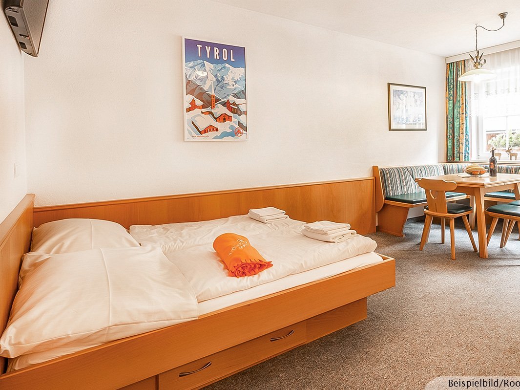 Das Bild zeigt ein Doppelbett mit Bettwäsche, Bilder an der Wand, Essecke mit Tisch, Eckbank und Stühlen