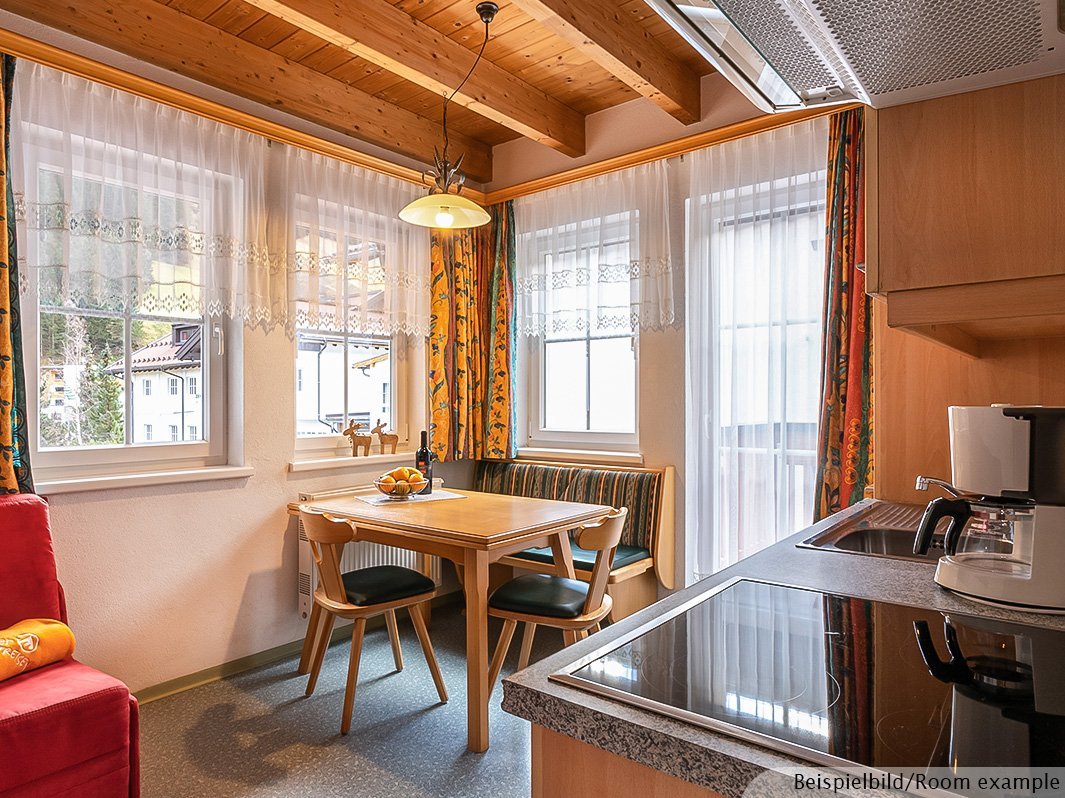 Das Bild zeigt Wohnzimmer mit Sitzecke, Tisch, Bank und Stühlen, Fenster, Tür zum Balkon, rotes Sofa, Küchenzeile mit Kaffeemaschine, Wasserkocher, Waschbecken, Herdplatte 