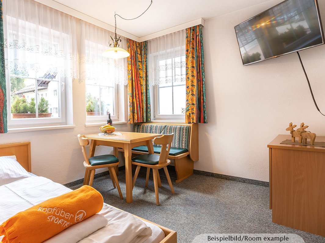 Das Bild zeigt ein Einzelbett mit Bettwäsche, Essecke mit Tisch, Eckbank und Stühlen, Kommode, Fernseher, Fenster